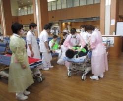 病院の待合室で白衣を着た医療者がストレッチャーに横たわっている男性の首元に手を当て訓練を行っている写真