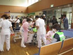 病院の待合室で、ストレッチャーや車いすに乗った女性、長椅子に座っている女性にトリアージを行っている看護師の人達の写真