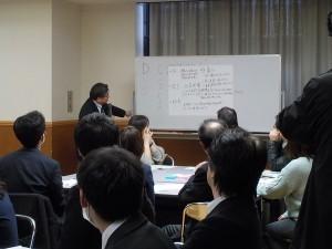 ホワイトボードに大きな模造紙を貼っている左側の男性の講義を、椅子に座って聞いている参加者の後ろ姿の写真