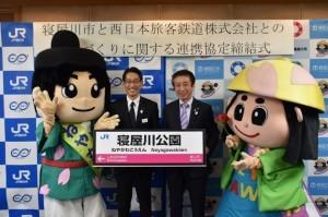 はちかづきちゃんとねや丸くんにの間に西日本旅客鉄道株式会社の関係者の男性と市長が寝屋川公園駅プレートを持って並んでいる協定書締結式での写真