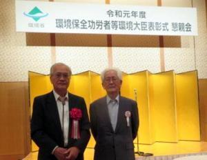 「令和元年度環境保全功労者等環境大臣表彰式と掲げられた吊り看板のある表彰式会場で2名の年配の男性が並んで写っている写真