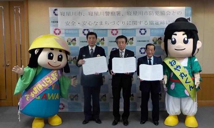 安全・安心なまちづくりに関する協定締結式にて、協定書を手に持ち、寝屋川市、寝屋川警察署、寝屋川市防犯協会の各代表者とはちかづきちゃん、ねや丸くんが並んで写っている協定締結式の写真