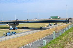 公園内の広場に橋梁がかかり、その上を自動車が走行しており、広場横では工事が行われている写真