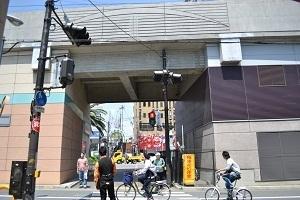 正面に大きな高架線があり、建物の手前にある横断歩道の信号機で信号待ちをしている方や、その前を自転車が通過しようとしている写真