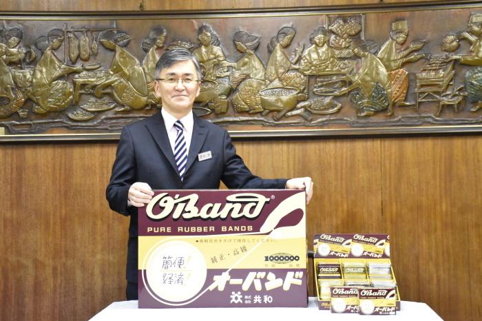 茶色と黄色のデザインのオーバンドパッケージを持って笑顔で写っている杉原正博さんの写真