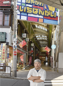 ねやがわいちばんがいの看板が掲げられている商店街の前で腕を組んでいる最東さんの写真