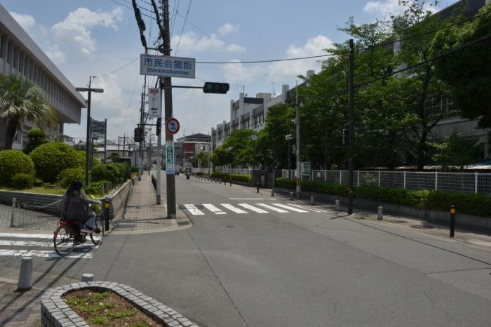 交差点の信号機が設置された電信柱に「市民会館前」と書かれた標識看板があり、道路の両脇に大きなコンクリートの建物が建っている写真