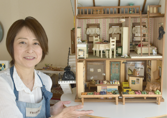 エプロン姿の河原かゆりさんと1階がお店、2階がダイニングルーム、寝室、ロフトになっているドールハウスの写真