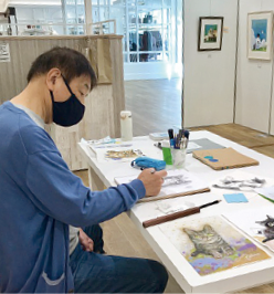 展示会場の机の上で作品作りに取り組んでいる勝間としをさんの写真