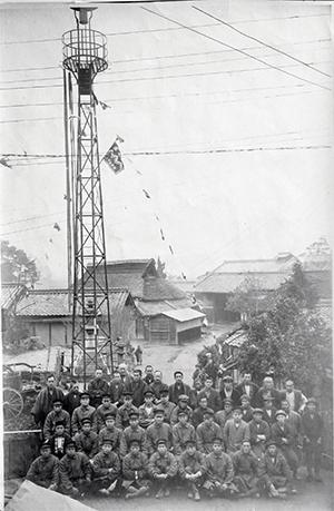 かやぶき屋根の民家が建ち並び、火の見やぐらの下で記念撮影している白黒写真