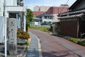 舗装された道路の左側に白い建物と記念碑が建ち、コンクリートの側溝にグレーチングが使用され、右側には木造の家が建っている写真