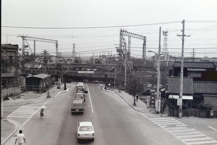 信号機のない道路に車が縦一列になり、踏切で止まっている白黒写真