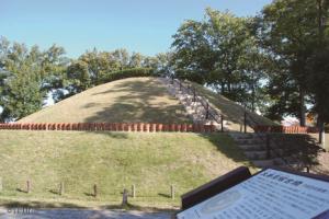 手前に古墳の説明看板、その奥に山のように盛り上がり、上り下りするための階段が設置された太秦高塚古墳が写っている写真