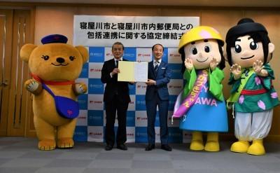 寝屋川市と寝屋川市内郵便局との包括連携に関する協定締結式と書かれた横断幕をバックに、ぽすくま、はちかづきちゃん・ねや丸くんのゆるキャラとの間に市長と郵便局の関係者の男性が横一列に立ち協定書を一緒に持っている写真
