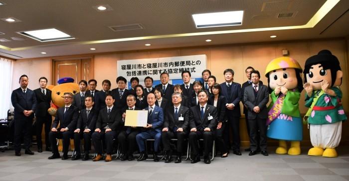 寝屋川市と寝屋川市内郵便局との包括連携に関する協定締結式と書かれた横断幕をバックに、ぽすくま 、はちかづきちゃん、ねや丸くんのゆるキャラと関係者が集まり、前方に7名の男性がパイプ椅子に座って、中央の2名の男性が一緒に協定書を持っている集合写真