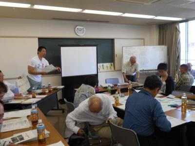 立って男性が資料を手に挨拶および素案の説明をするのを聞いている方々の写真