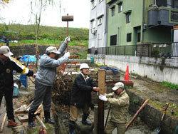 コンクリートを流した大きな溝の中央に立てたくいを2名の男性が支え、溝の上から木槌を持った男性が振り上げて上から打とうとしている様子の写真