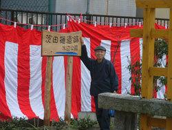 公園を隔てた柵沿いに大きな紅白幕を掛けた前で、男性が「茨田樋遺跡水辺公園」と書かれた手作りの木製の看板に右手を添えている写真
