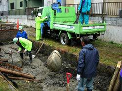 トラックから吊り下げたワイヤーで縛った大きな石を、川の底に置いている様子の写真