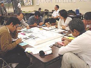 机を囲んで座り、それぞれ手元にある紙に描き込んでいる参加者の写真