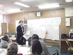 資料が貼られているホワイトボードの左側にスーツを着た男性が立ち説明しているのを、参加者が聞いている写真