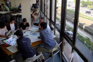 テーブルの上に資料を広げている参加者が座る右側に、寝屋川を見下ろせる大きなガラス窓が設置されているワークショップ会場の写真