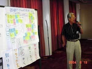 左側のホワイトボードに大きな模造紙が貼られており、右側に立つ本多会長が左手にマイクを持ち話しをしている写真