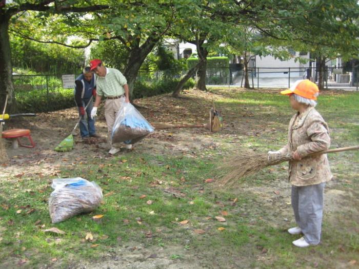 公園で落ち葉を集めたごみ袋を持っている赤い帽子を被った男性と、熊手で落ち葉を集めている青いベストを着用した男性の方を帽子を被り箒を持った女性が見ている写真