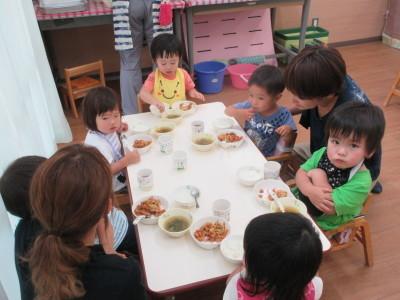 机の上に置かれた給食を子供たちが食べていて、お母さん2人がその様子を見ている写真