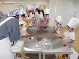 調理室の作業台にエプロンと三角巾を付けた子ども達や女性が、お団子のようなものを丸めている写真