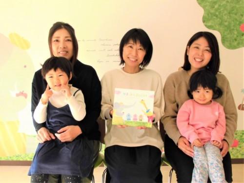 中央に絵本を持った女性、両脇に子供を膝の上に乗せ笑顔で座っている女性が写っている写真