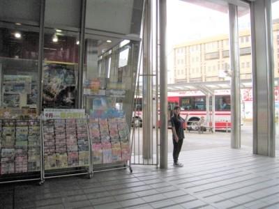 旅行のパンフレットが沢山並んで置かれてある店舗の横に立っている女性と、女性の奥にバスロータリーが見えている写真