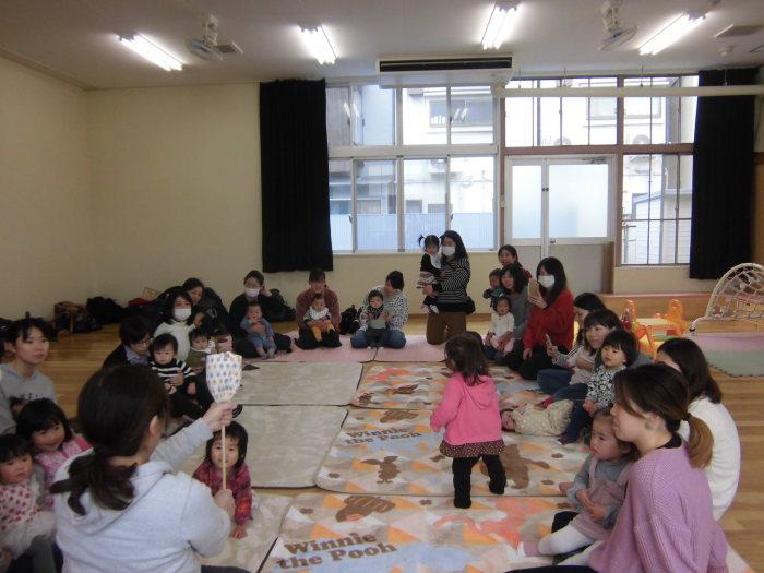 教室内に敷いたマットの上で円を描くようにまるくなって座っている親子連れの写真