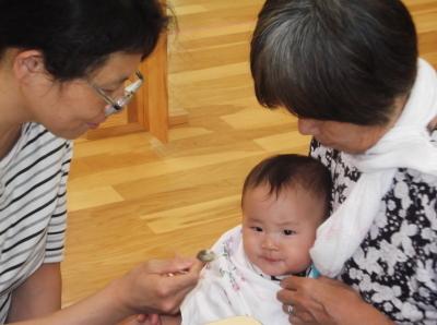 おばあちゃんに抱かれている赤ちゃんと赤ちゃんに離乳食を食べさせようとしている母親の写真