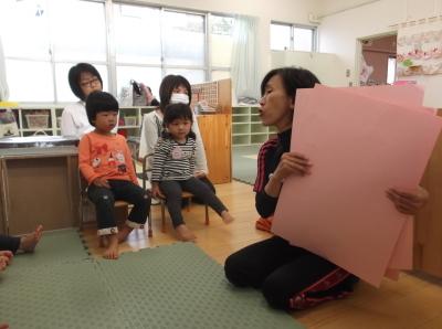 ピンク色の画用紙を持っている先生、椅子に座っている子供とその後ろに座っている母親の写真