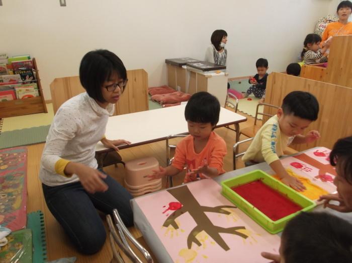 赤や黄色の絵の具を手のひらにつけて木の枝の形の画用紙に手形をつけている子供たちの写真