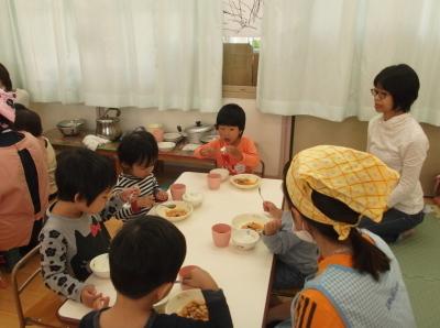 席について給食っを食べている子供たち、テーブルの近くでその様子を見ている母親と先生の写真