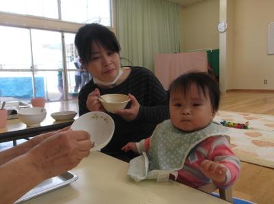 前掛けをして椅子に座っている赤ちゃんと横で茶碗とスプーンをもって離乳食を食べさせている母親の写真