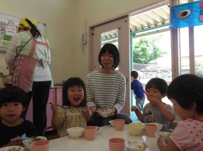 3歳児クラスの子供たちと一緒に給食を食べている女の子と母親、その後ろに立っている先生の写真