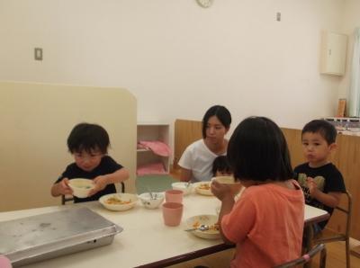 席について給食を食べている3人の子供たちと赤ちゃんを膝に抱いて座っている母親の写真