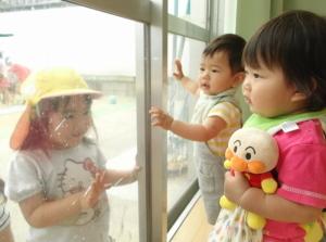 窓の外から中を見ている黄色い帽子の女の子、アンパンマンのぬいぐるみを持っている子供と窓に手をかけて立っている子供の写真