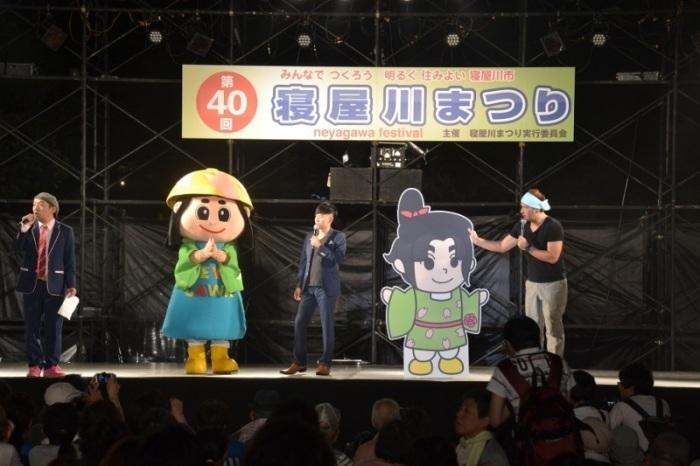 舞台上に、はちかづきちゃんとねや丸くんの等身大パネルとマイクを持った男性3名がイベントを行っている第40回寝屋川まつりの写真