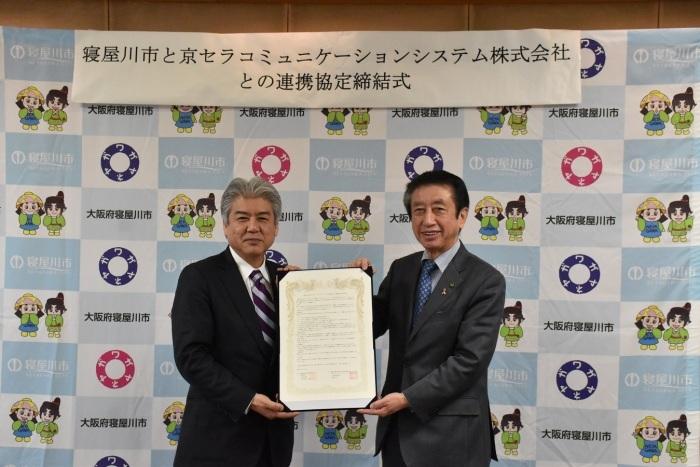 寝屋川市と京セラコミュニケーションシステム株式会社との連携協定締結式と書かれた横長の貼り紙の前で、2名の男性が締結書を一緒に両手で持っている写真