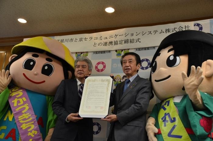2名の男性が一緒に両手で締結書を持っており、両側にゆるキャラ1体ずつと並んだ集合写真