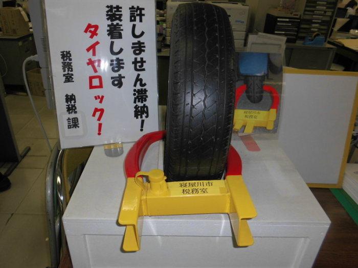 「許しません滞納！装着しますタイヤロック！税務室納税課」と書かれた貼り紙の右側に、タイヤロックされたタイヤが事務所の白い台の上に置かれている写真