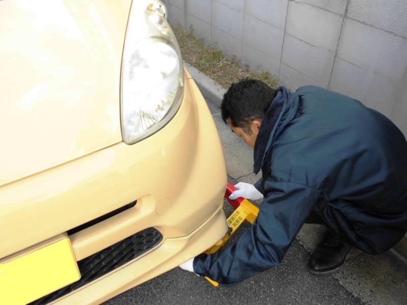 紺色のジャンパーを着た男性が、薄い黄色の車の右前のタイヤにタイヤロックを行っている写真