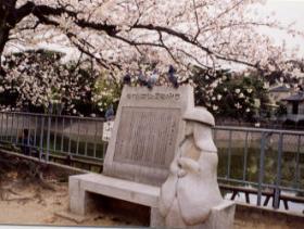 桜が咲いている桜の木の側に鉢かつぎ姫の形をした石像と説明看板が立てられている写真