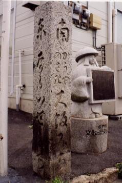 四ツ辻に立つ道標と隣に鉢かつぎ姫の形をした石像が説明看板を持っている写真