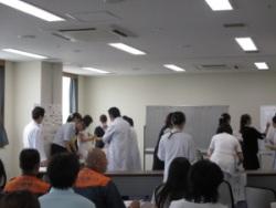 会議室前方にあるホワイトボードに記入したり、白衣を着用している医療者の人達が訓練を行っているのを椅子に座って見ている参加者の後ろ姿の写真