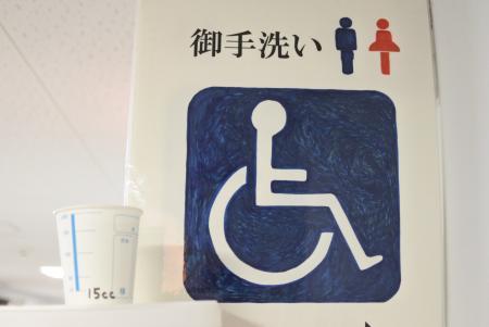 横書きで御手洗いと書かれた文字の横にトイレの男女のマークと下に車いす用のマークがあり、左側の棚に検尿カップが置かれている写真